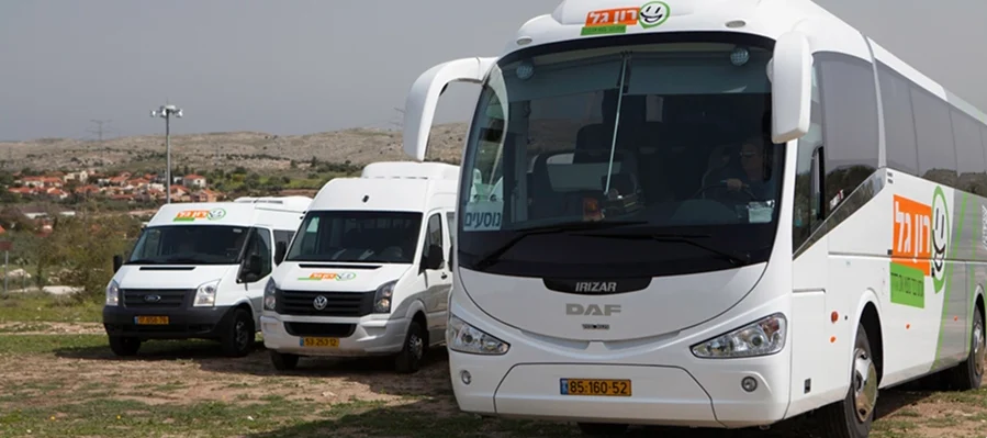 שלוש מכוניות נוסעים לבנות, כולל אוטובוס תיירים ושני טנדרים, חונות בחוץ על שטח עפר, כשברקע נראים גבעות ובתים.