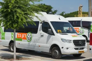 מיניבוס לבן של מרצדס-בנץ עם כיתוב בעברית בצבעים ירוק וכתום, חונה בחניון ליד עץ; ברקע נראה רכב דומה נוסף.
